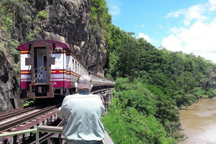 A train on tracks, a man standing in front of it, and a river beside a forested cliff. 1