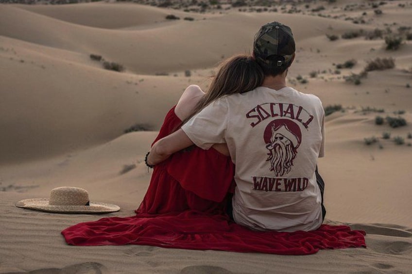 Twee personen zitten op een kleed in een woestijn, met zandduinen op de achtergrond. 5