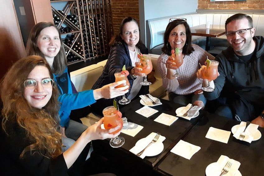 A group of people sitting at a table with drinks, in a restaurant with a brick wall and wine racks in the background. 2