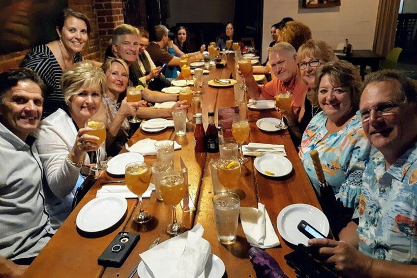 A group of people seated around a long wooden table with drinks and plates. 3