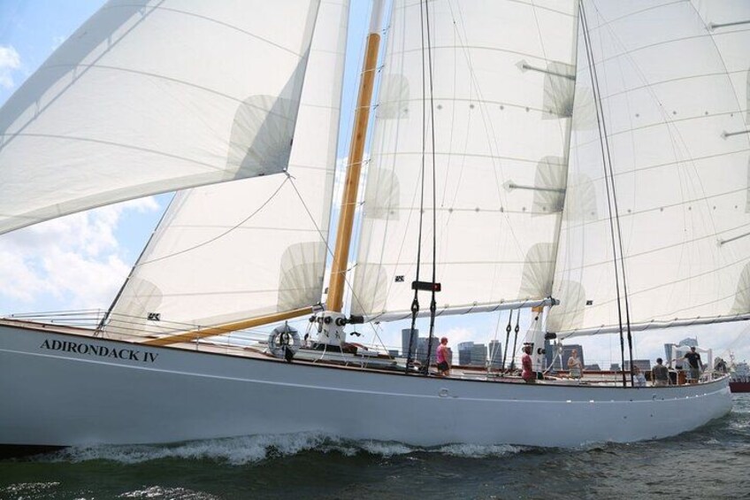 Een zeilboot genaamd 'ADIRONDACK IV' met meerdere zeilen, mensen aan dek en een stadshemelbeeld op de achtergrond. 3