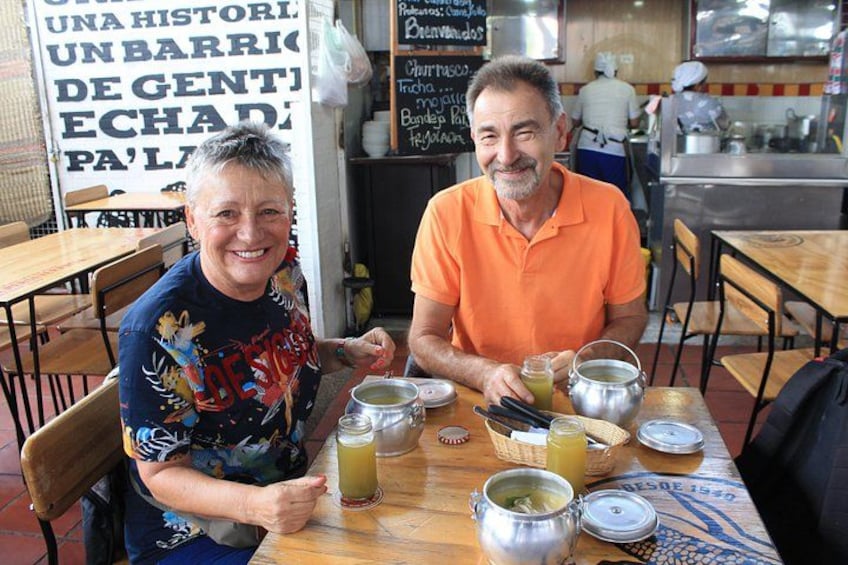 Twee personen zitten aan een houten tafel met verschillende soorten eten en drinken. 4