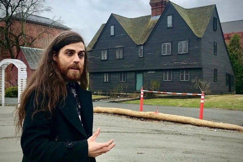 A person standing in front of a large, dark-colored house with a steep roof and multiple windows. 1