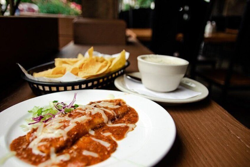 A plate of enchiladas with sauce, a bowl of chips, and a cup of coffee on a wooden table. 5