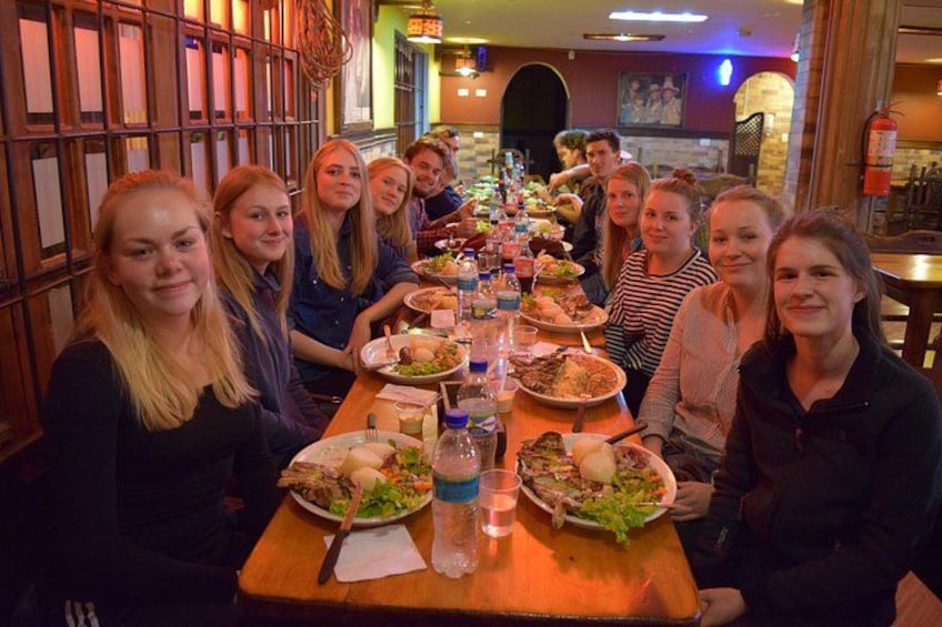 Een groep mensen zit rond een lange houten tafel met verschillende gerechten en drankjes. 5