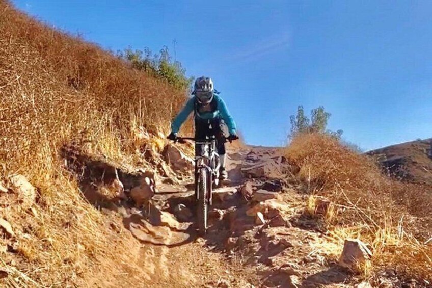 A person riding a mountain bike on a rocky trail surrounded by dry grass and hills. 1