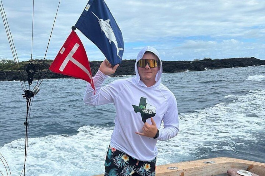 Une personne à bord d’un bateau, tenant un drapeau orné d’un motif de requin, portant un sweat à capuche blanc et un short à motifs floraux. 3