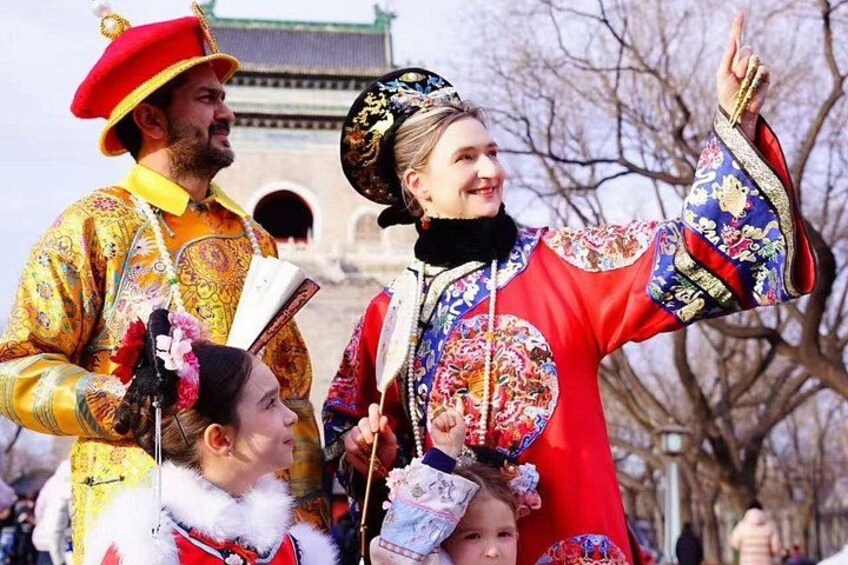 A person in traditional red and blue attire with floral patterns, holding a fan, and standing in front of a building with a traditional roof. 1