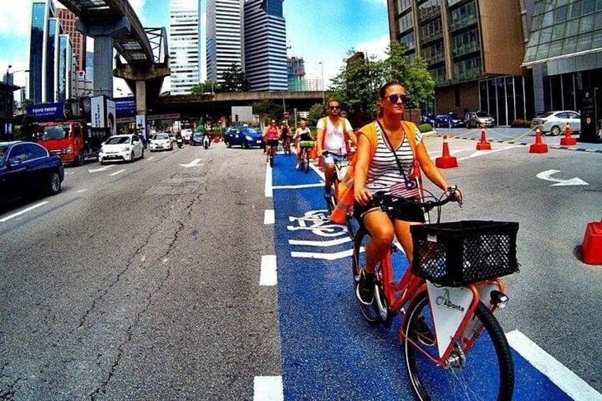 Un cycliste sur une piste cyclable bleue, avec un panier sur son vélo, une voiture et un piéton en arrière-plan. 2