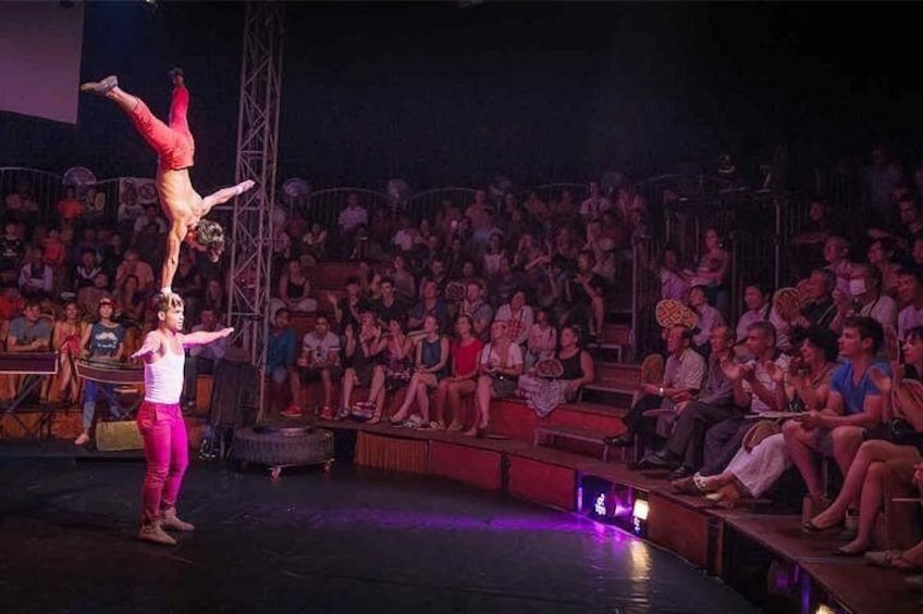 En cirkusföreställning med akrobater på scenen, en publik som sitter längre bak och en scen med en metallkonstruktion. 4