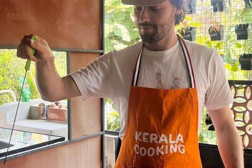 Une personne portant un tablier sur lequel est écrit « CUISINE DU KERALA », tenant un objet vert, se tient à l’intérieur, près d’une fenêtre, avec des plantes en arrière-plan. 3