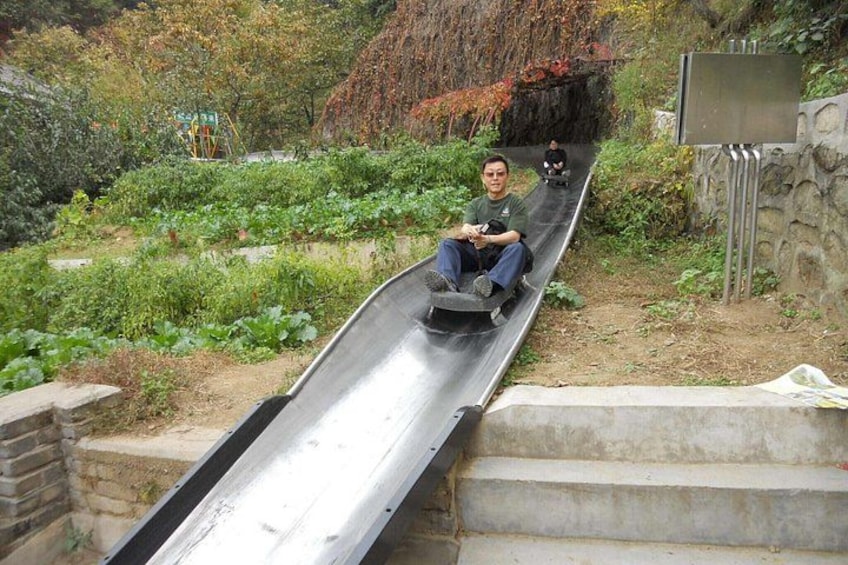 A person sliding down a metal slide in a garden with vegetation and a stone wall. 3