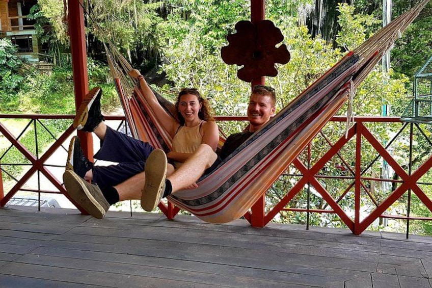 Two people relaxing in a hammock on a wooden deck with a red railing and greenery in the background. 2