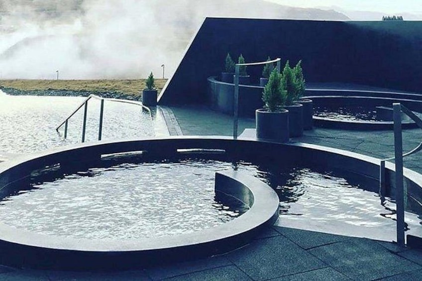 Una estructura arquitectónica moderna con un elemento acuático circular, plantas en macetas y vistas a un paisaje lejano. 3