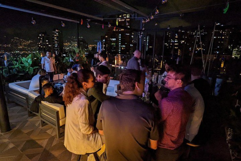 A rooftop bar with people socializing, a city skyline in the background, and a view of the city. 3