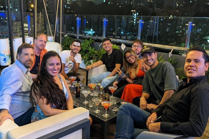 A group of people seated around a table with drinks and glasses, in a room with large windows overlooking a cityscape. 5