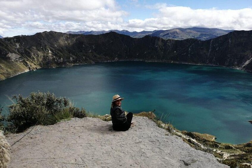 A person sitting on a rocky ledge overlooking a large body of water surrounded by mountains. 1