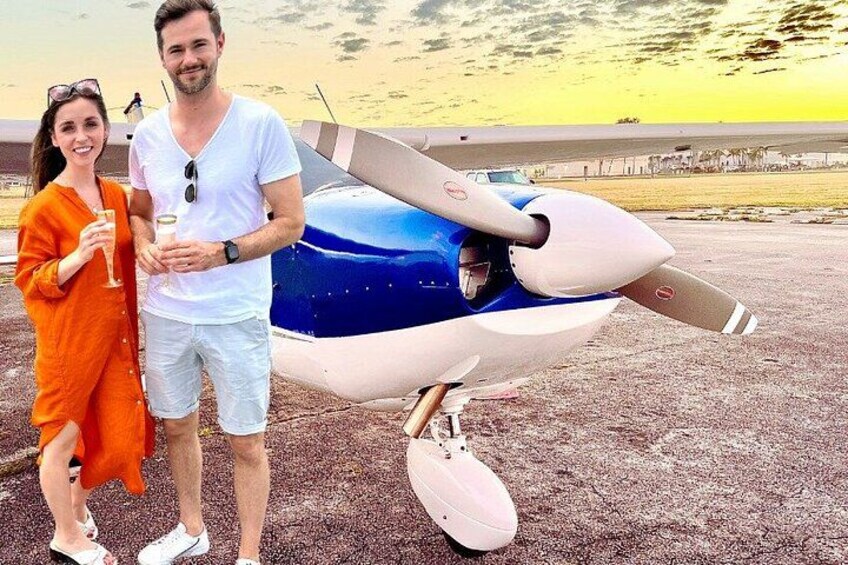 Un couple se tient à côté d’un petit avion bleu et blanc sur une piste, avec un coucher de soleil en arrière-plan. 4