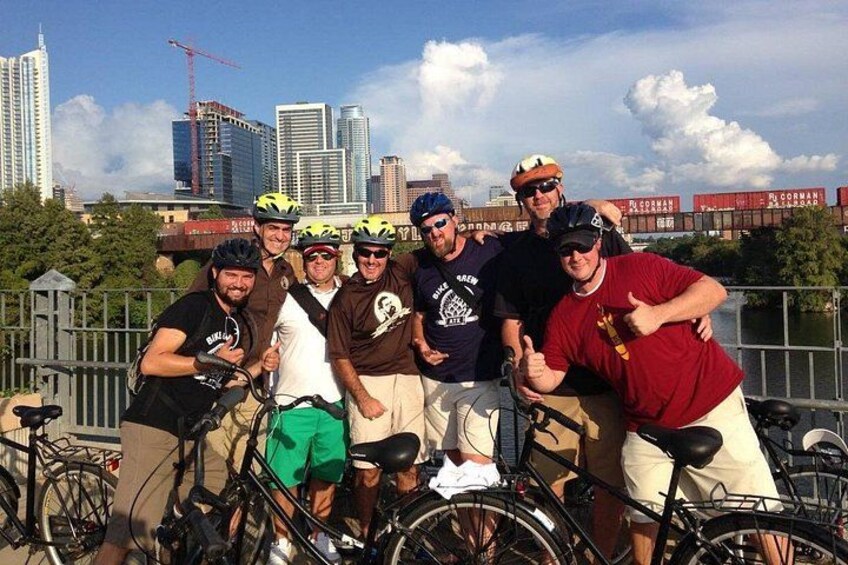 A group of cyclists posing with bicycles in an urban setting with a river and skyscrapers in the background. 1