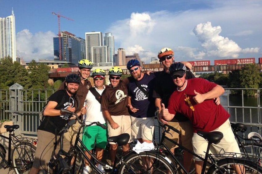 A group of cyclists posing with their bikes in an urban setting with a city skyline and construction cranes in the background. 1
