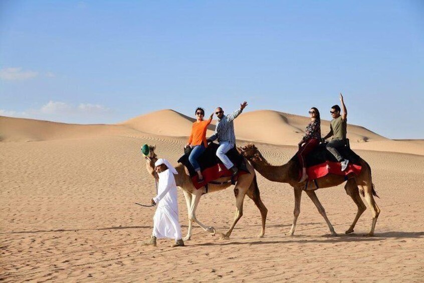 Eine Gruppe von Menschen reitet auf Kamelen durch eine Wüstenlandschaft mit Sanddünen im Hintergrund. 3