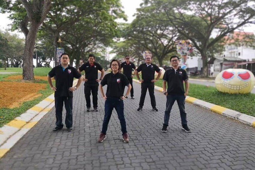 A group of people in black shirts standing on a paved path in a park with trees and a large yellow sculpture in the background. 1