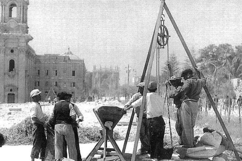 Eine historische Bautätigkeit mit Arbeitern, die einen Kran und ein Seilwindensystem verwenden, einer Kirche im Hintergrund und einer ländlichen Landschaft. 2