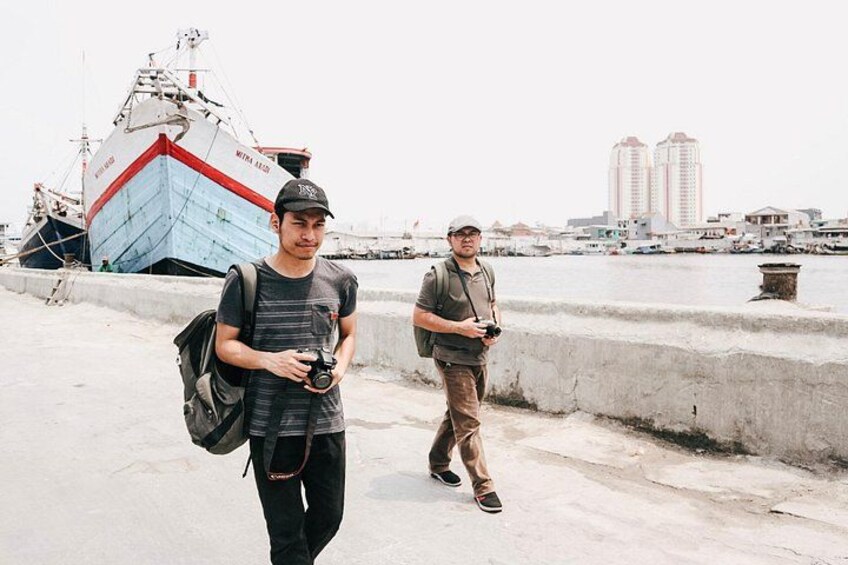 Dos personas caminando cerca de un muelle, con un barco grande y edificios al fondo. 2