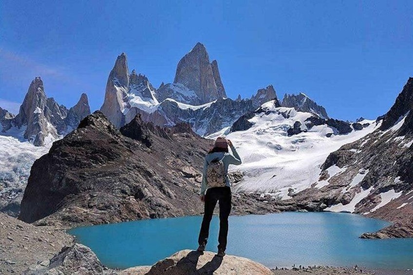 A person standing on a rocky outcrop with a lake and snow-capped mountains in the background. 1