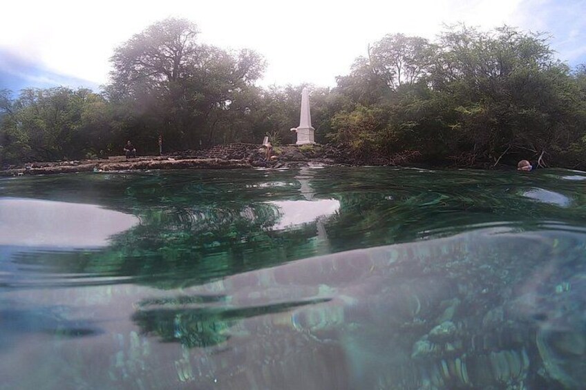 A clear body of water with a statue in the background and dense foliage around. 3
