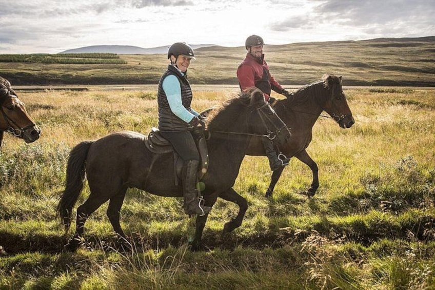 Two people riding horses in a grassy field with rolling hills in the background. 1