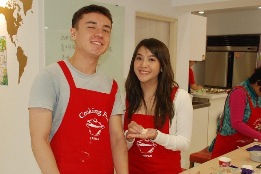 Two people wearing red aprons with a cooking logo, standing in a kitchen with a stainless steel refrigerator and a whiteboard in the background. 5