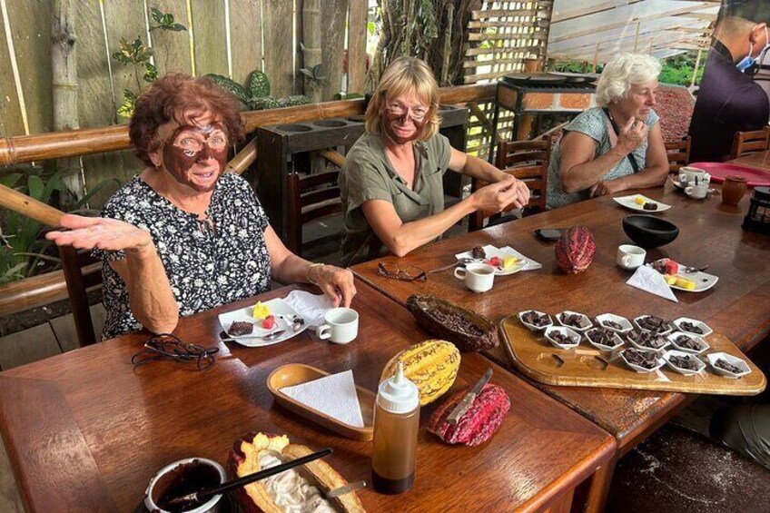 A group of people sitting at a wooden table in an outdoor setting, enjoying a meal with various dishes and drinks. 4