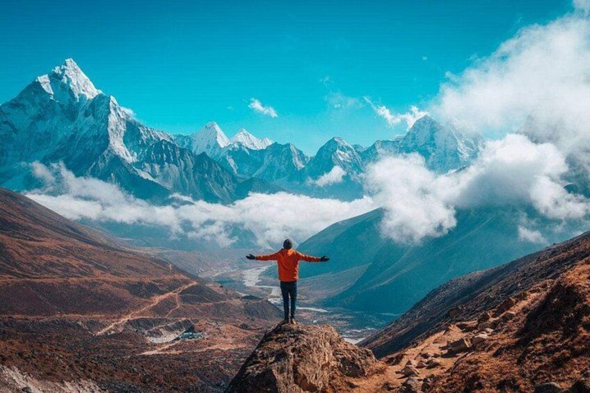 Una persona de pie sobre una cresta montañosa con los brazos extendidos, contemplando un valle con montañas nevadas y nubes abajo. 5