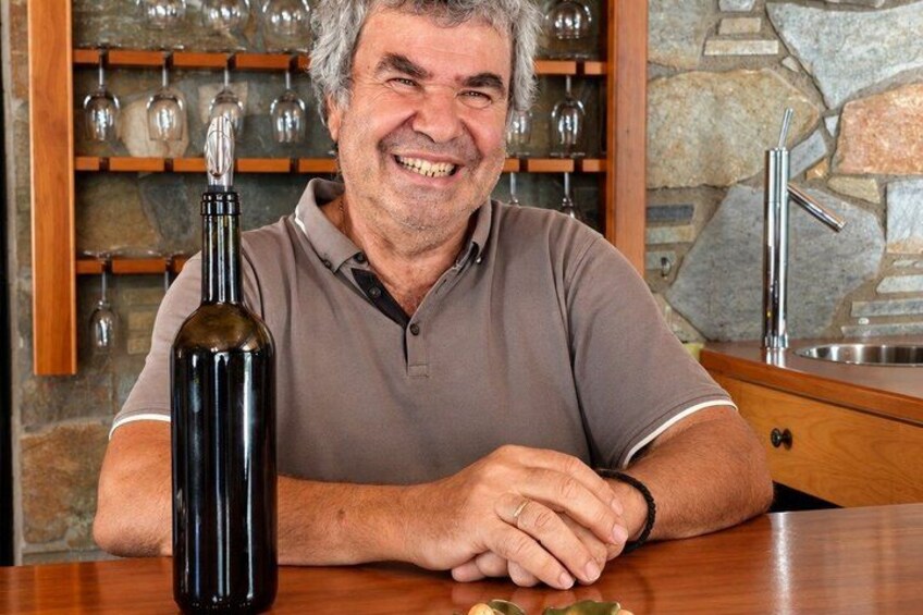 Un homme est assis à une table devant laquelle se trouve une bouteille de vin, un évier de cuisine et un mur en pierre orné d’étagères où sont posés des verres à vin. 4