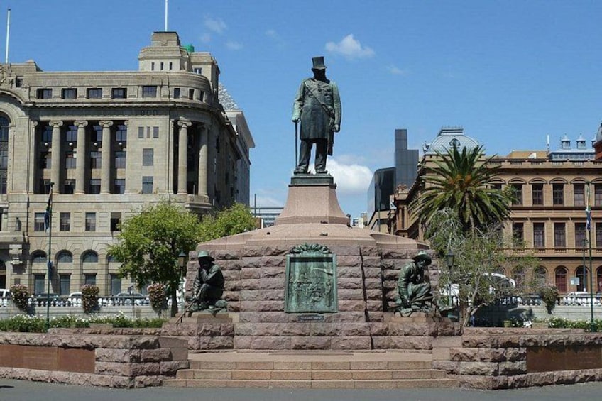 A statue of a man in uniform on a pedestal with two smaller statues on either side, set against a backdrop of a large building and a palm tree. 1