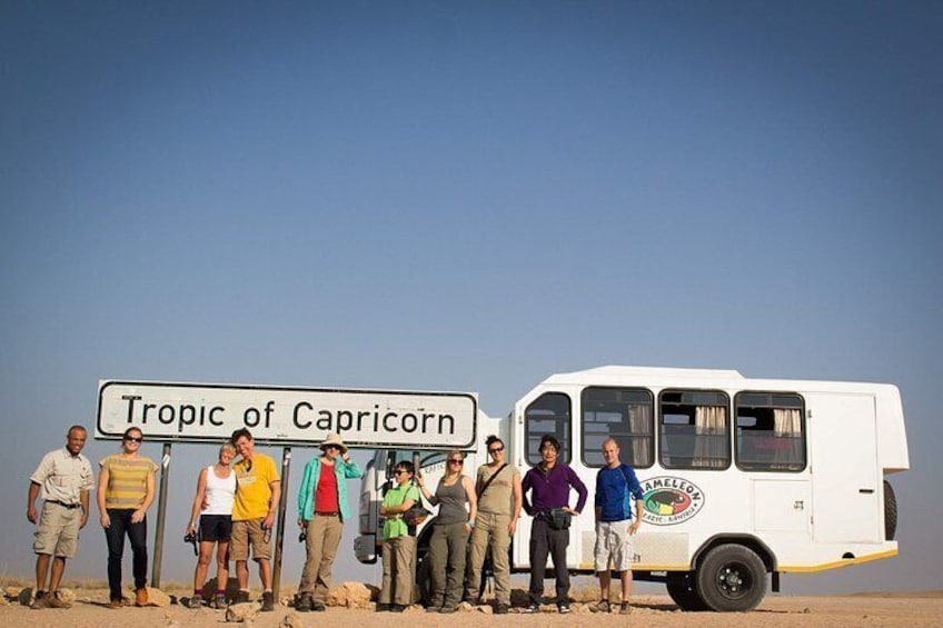 A group of people standing in a desert with a sign that reads 'Tropic of Capricorn' and a white van with the logo 'Amelco Music Adventures.' 4