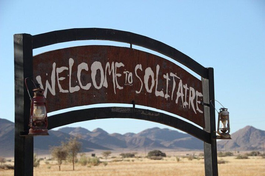 A rustic welcome sign with 'WELCOME TO SOLITAIRE' written on it, two lanterns, and a desert landscape with mountains in the background. 5