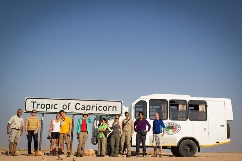 A group of people standing in a desert with a sign that reads 'Tropic of Capricorn' and a white van. 2