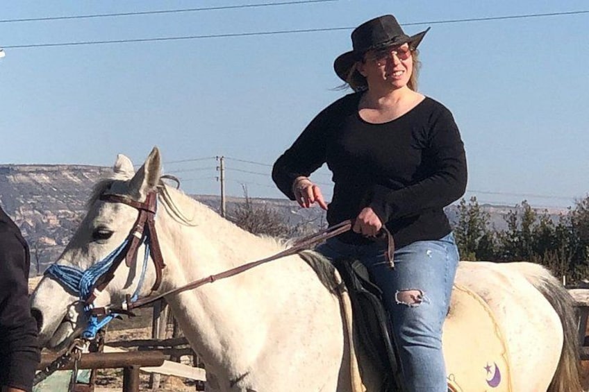 A person riding a white horse with a blue halter, wearing a black shirt and jeans, in an outdoor setting with a clear sky. 3