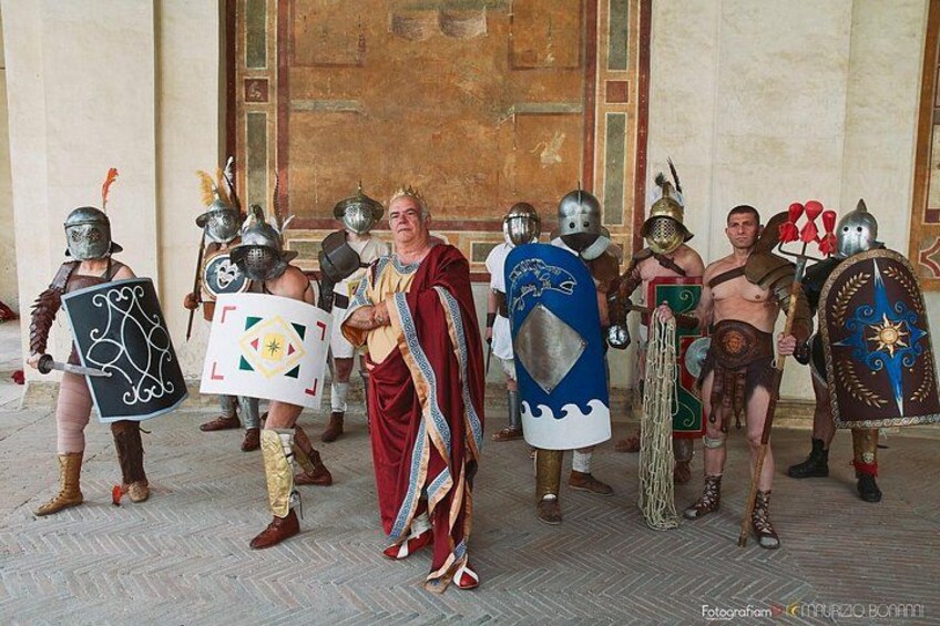 Un grupo de personas vestidas con trajes y armaduras históricas, sosteniendo escudos y espadas, de pie en una habitación con una gran puerta de madera al fondo. 3