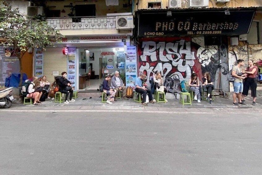 A street with people sitting on stools, a barber shop, and a pizza shop. 3