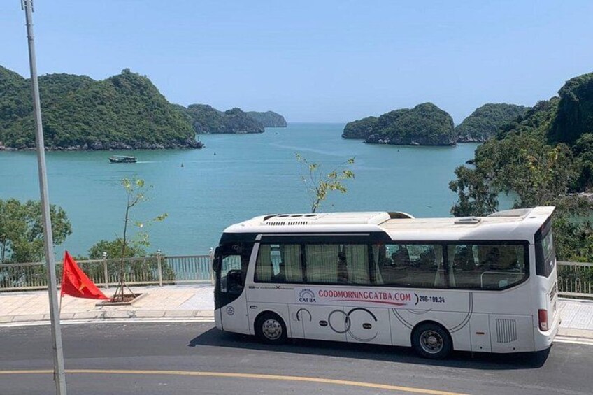 A white bus with a website URL on its side, parked on a road with a view of a coastal area and hills. 4
