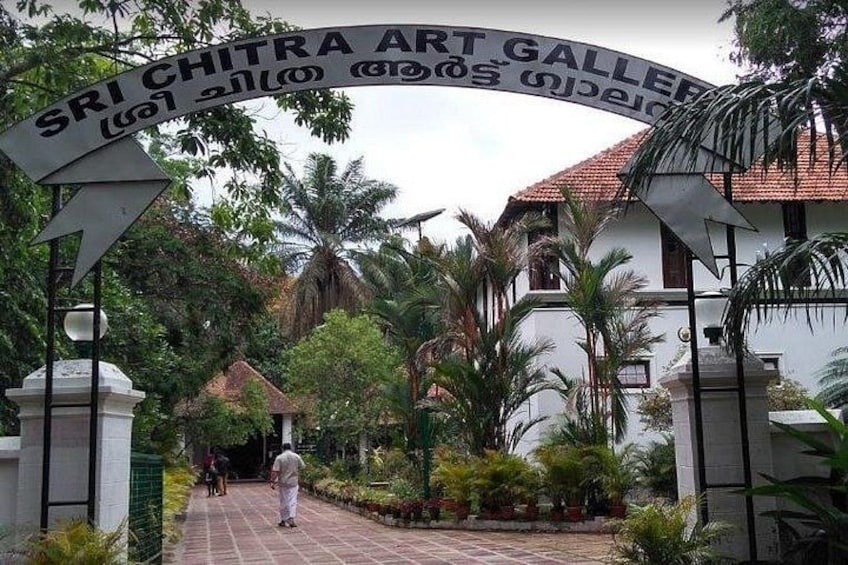 La entrada a la Galería de Arte Sri Chitra, con un letrero en inglés y otro idioma, rodeada de exuberante vegetación y palmeras. 2