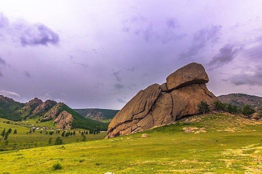A large rock formation in a grassy field with mountains in the background. 1