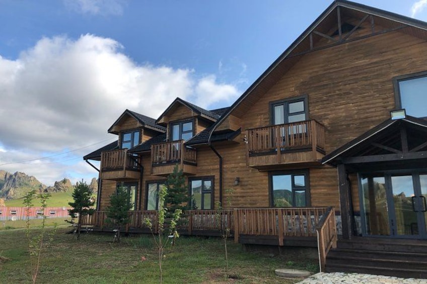 A wooden multi-story building with balconies, surrounded by a grassy area and trees. 3