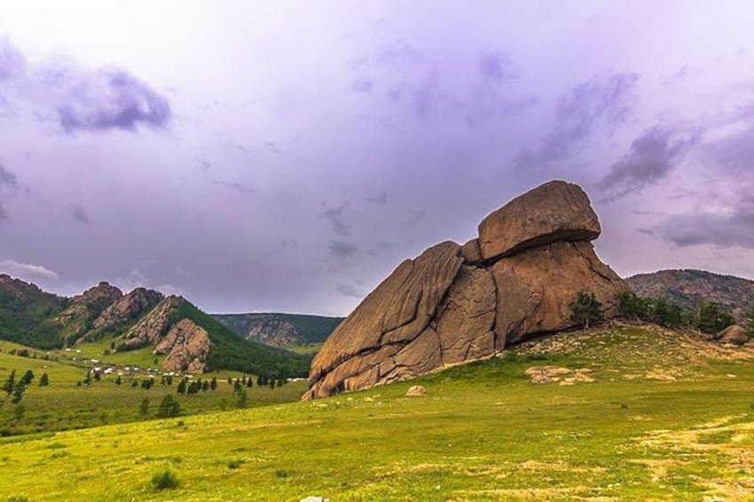 A large rock formation in a grassy field with mountains in the background. 1