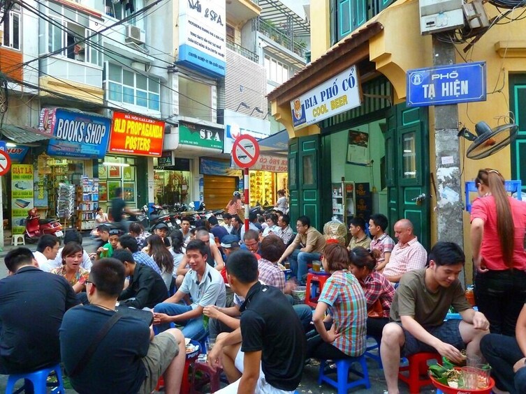 A crowded street with people sitting on stools, a books shop, and a sign for 'BIA PHÒ CÓ cool beer'. 5
