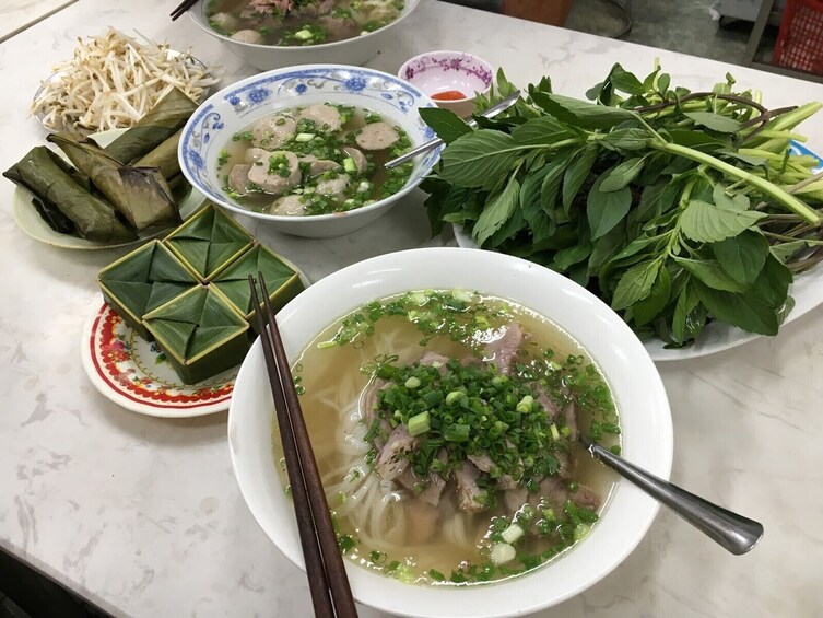 A table with various dishes including a bowl of soup with meat and green onions, a plate of bean sprouts, and several dishes wrapped in leaves. 2