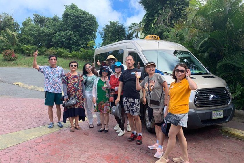 A group of people posing in front of a taxi with a 'TAXI' sign on a paved area with greenery in the background. 1